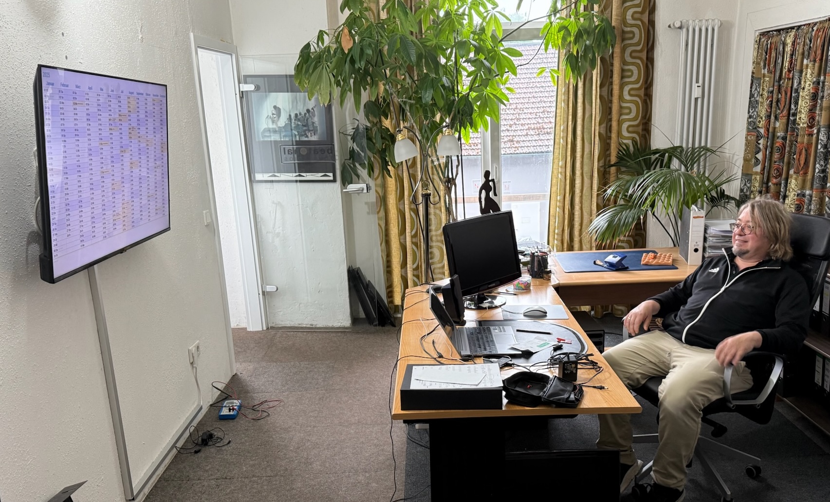 Piano teacher sitting at his desk, looking at the wall-mounted monitor displaying the calendar